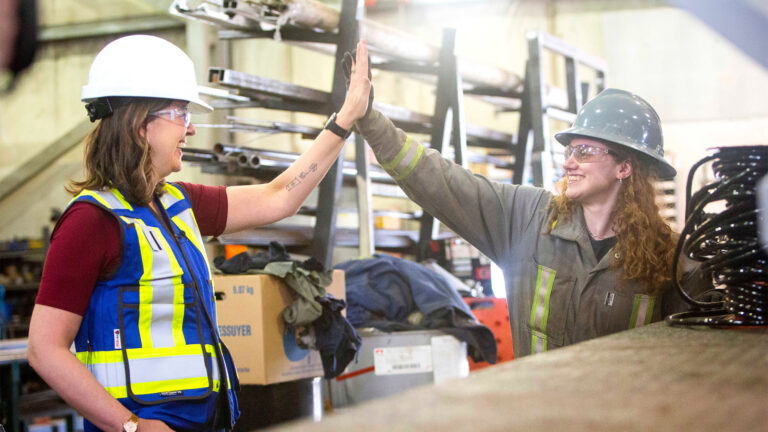 Danielle Smith in a hard hat sharing a high-five with a tradeswoman at an industrial plant