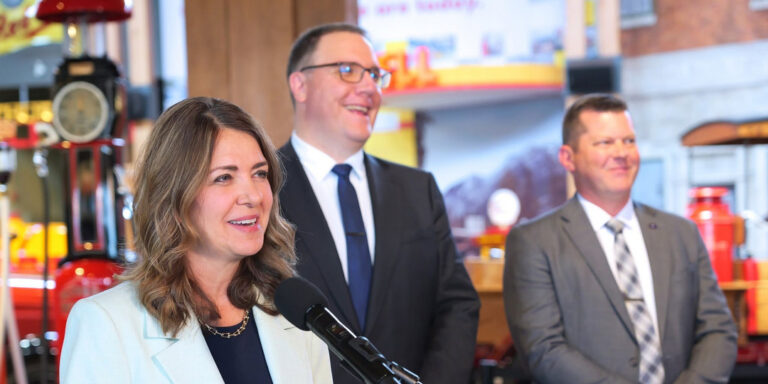 Danielle Smith speaking at a microphone with two colleagues behind her