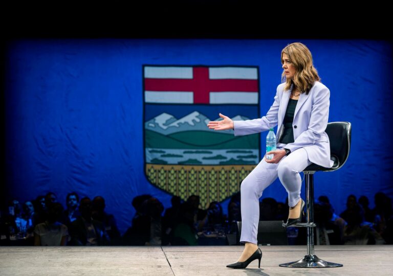 Danielle Smith seated on stage beside the Alberta coat of arms